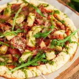 Pizza con masa de pizza sin gluten, cubierta con alcachofas, jamón y pesto, decorada con rúcula fresca.