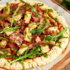 Pizza con masa de pizza sin gluten, cubierta con alcachofas, jamón y pesto, decorada con rúcula fresca.