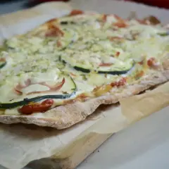 Pizza casera con base de pan, cubierta de queso derretido, rodajas de calabacín, jamón y tomates cherry.
