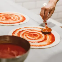 Masa de pizza casera lista para ser cubierta con salsa. Una mano con una cuchara extiende la salsa de tomate en espiral sobre la base.