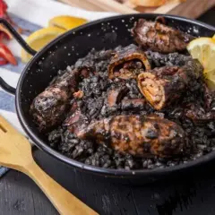Calamares rellenos en arroz negro, servidos en una paellera con gajos de limón y una cuchara de madera.