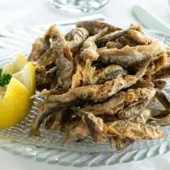 Deliciosa fritura de pescado crujiente servida en un plato de cristal con forma de pez, acompañada de un limón tallado y perejil fresco.