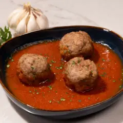 Tres albóndigas con tomate caseras, adornadas con perejil picado, junto a un ajo y perejil fresco.