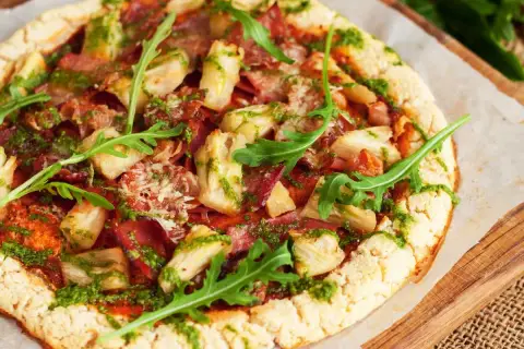 Pizza con masa de pizza sin gluten, cubierta con alcachofas, jamón y pesto, decorada con rúcula fresca.
