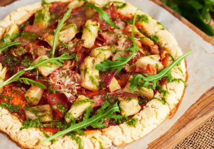 Pizza con masa de pizza sin gluten, cubierta con alcachofas, jamón y pesto, decorada con rúcula fresca.