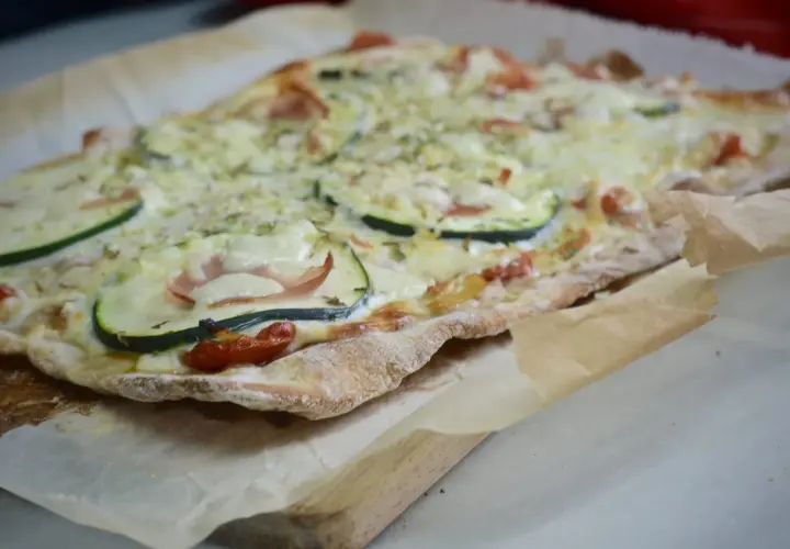 Pizza casera con base de pan, cubierta de queso derretido, rodajas de calabacín, jamón y tomates cherry.