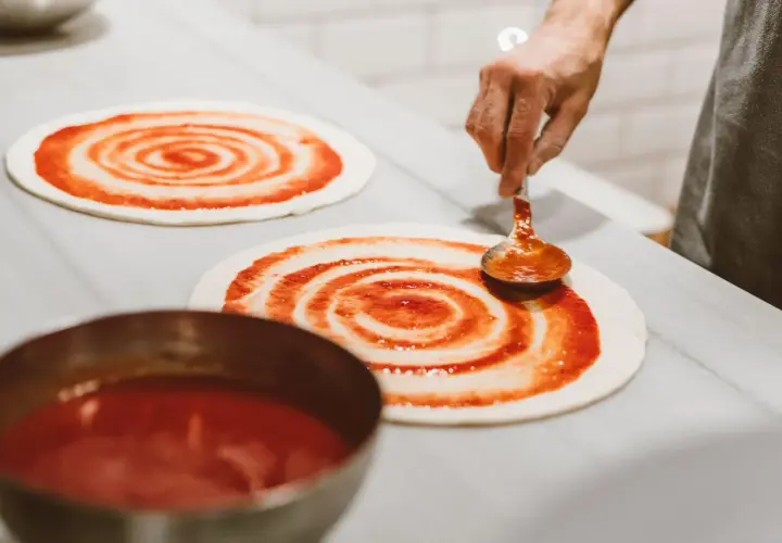 Masa de pizza casera lista para ser cubierta con salsa. Una mano con una cuchara extiende la salsa de tomate en espiral sobre la base.