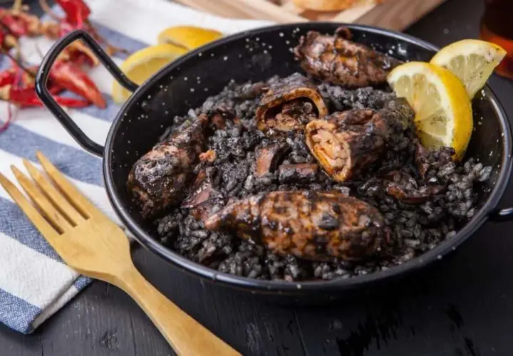 Calamares rellenos en arroz negro, servidos en una paellera con gajos de limón y una cuchara de madera.
