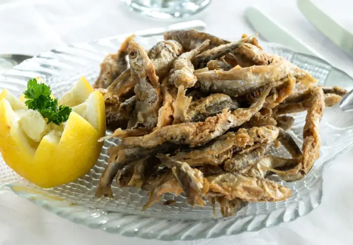 Deliciosa fritura de pescado crujiente servida en un plato de cristal con forma de pez, acompañada de un limón tallado y perejil fresco.