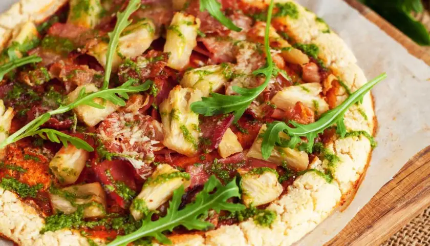 Pizza con masa de pizza sin gluten, cubierta con alcachofas, jamón y pesto, decorada con rúcula fresca.
