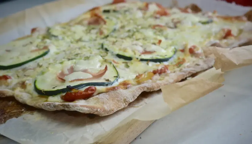 Pizza casera con base de pan, cubierta de queso derretido, rodajas de calabacín, jamón y tomates cherry.