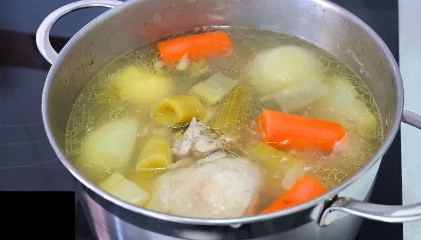 Olla con caldo de pollo casero hirviendo, con trozos de zanahoria, puerro, cebolla y pollo.