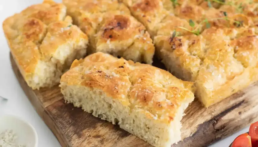 Focaccia dorada y tierna con sal gruesa y romero, cortada en porciones sobre tabla de madera.