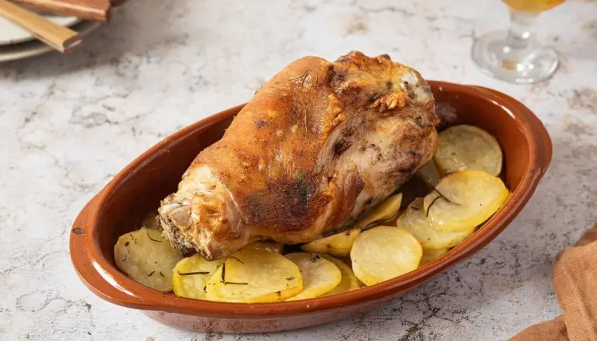 Codillo de cerdo asado, crujiente y dorado, servido en cazuela de barro con patatas panadera.