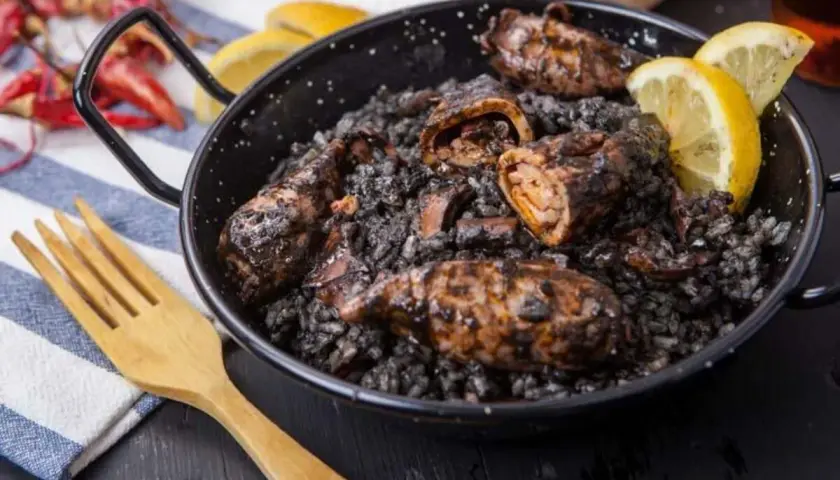 Calamares rellenos en arroz negro, servidos en una paellera con gajos de limón y una cuchara de madera.