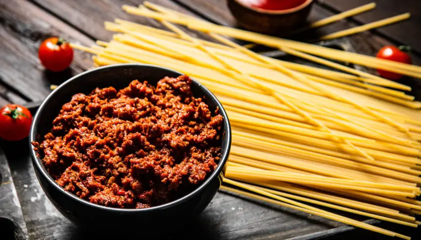Un tazón de carne boloñesa junto a espaguetis crudos y tomates. ¡Listo para unos deliciosos espaguetis a la boloñesa!