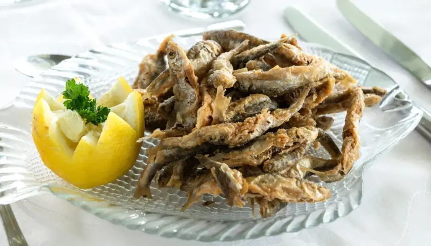 Deliciosa fritura de pescado crujiente servida en un plato de cristal con forma de pez, acompañada de un limón tallado y perejil fresco.