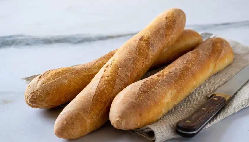 Tres baguettes doradas recién horneadas, listas para aprender como hacer baguette. Un cuchillo de madera descansa a su lado.
