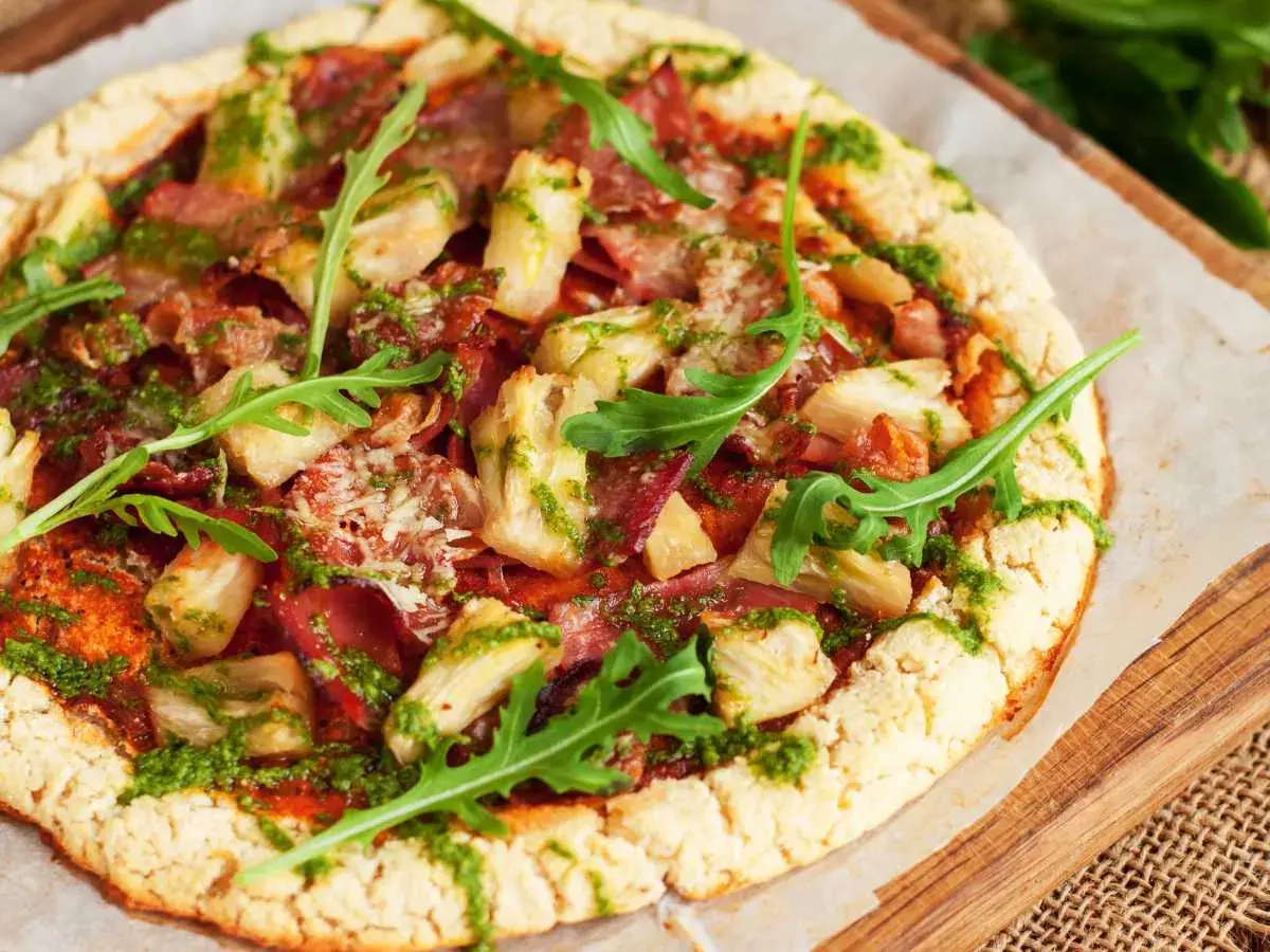 Pizza con masa de pizza sin gluten, cubierta con alcachofas, jamón y pesto, decorada con rúcula fresca.