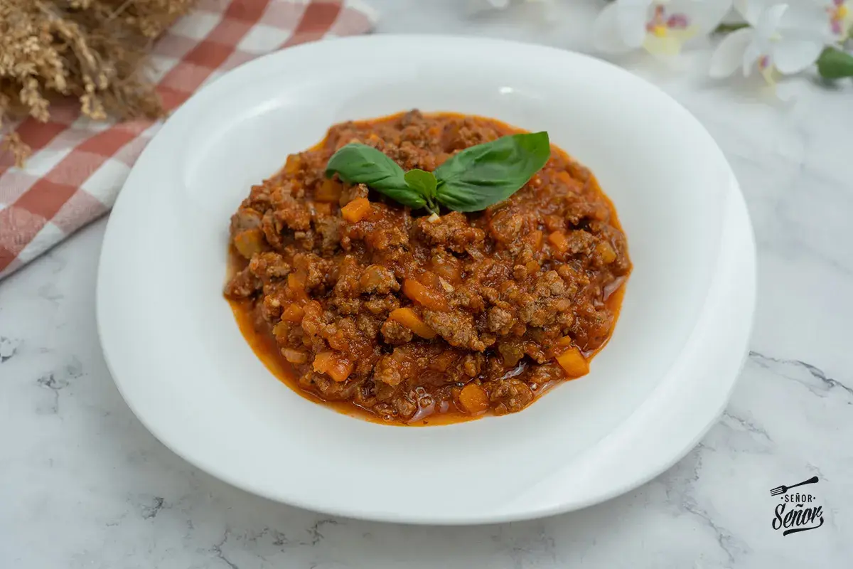 Un plato blanco con abundante salsa boloñesa casera, decorada con hojas de albahaca fresca.