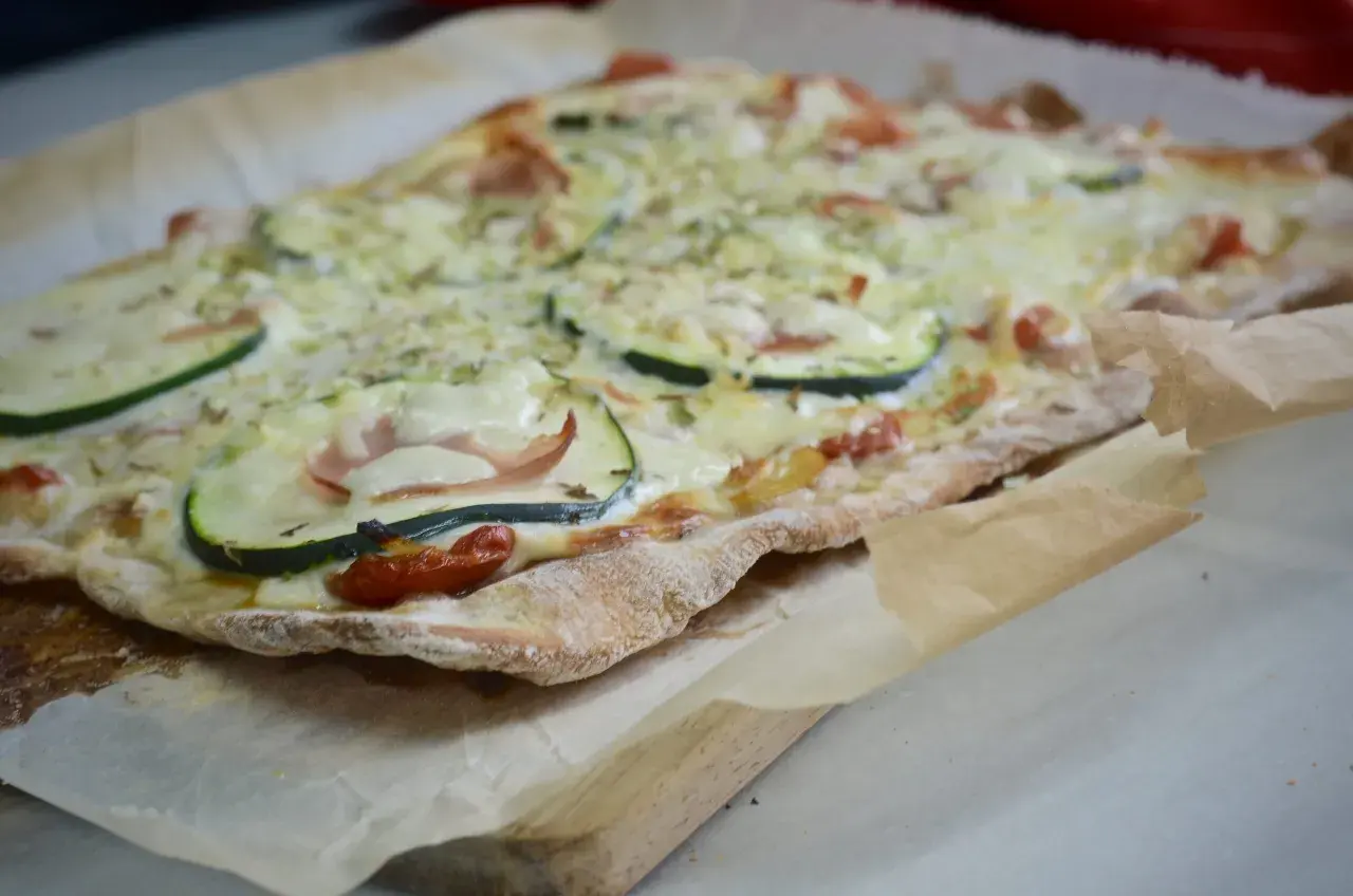 Pizza casera con base de pan, cubierta de queso derretido, rodajas de calabacín, jamón y tomates cherry.
