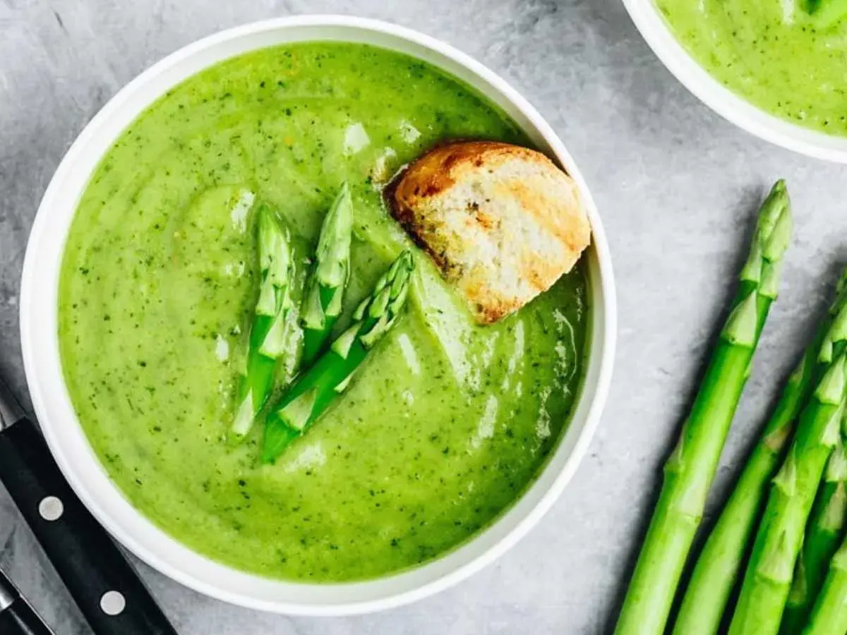 Crema de espárragos verde vibrante, decorada con espárragos frescos y un trozo de pan tostado.