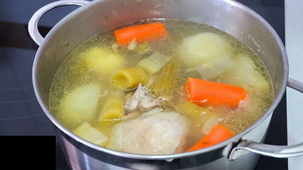 Olla con caldo de pollo casero hirviendo, con trozos de zanahoria, puerro, cebolla y pollo.