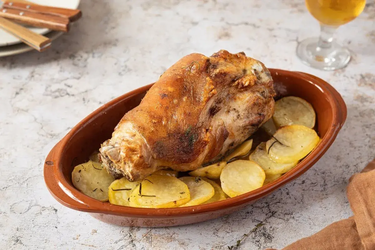 Codillo de cerdo asado, crujiente y dorado, servido en cazuela de barro con patatas panadera.