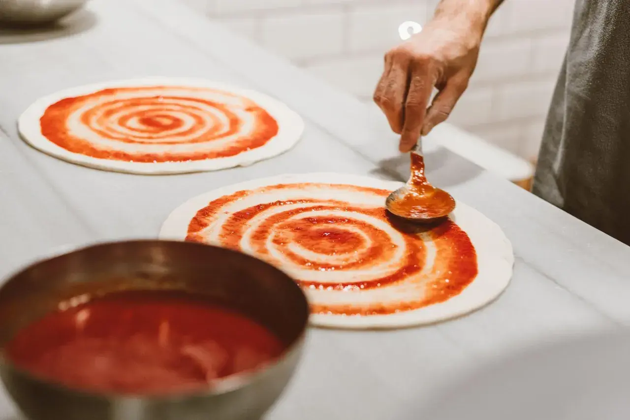Masa de pizza casera lista para ser cubierta con salsa. Una mano con una cuchara extiende la salsa de tomate en espiral sobre la base.