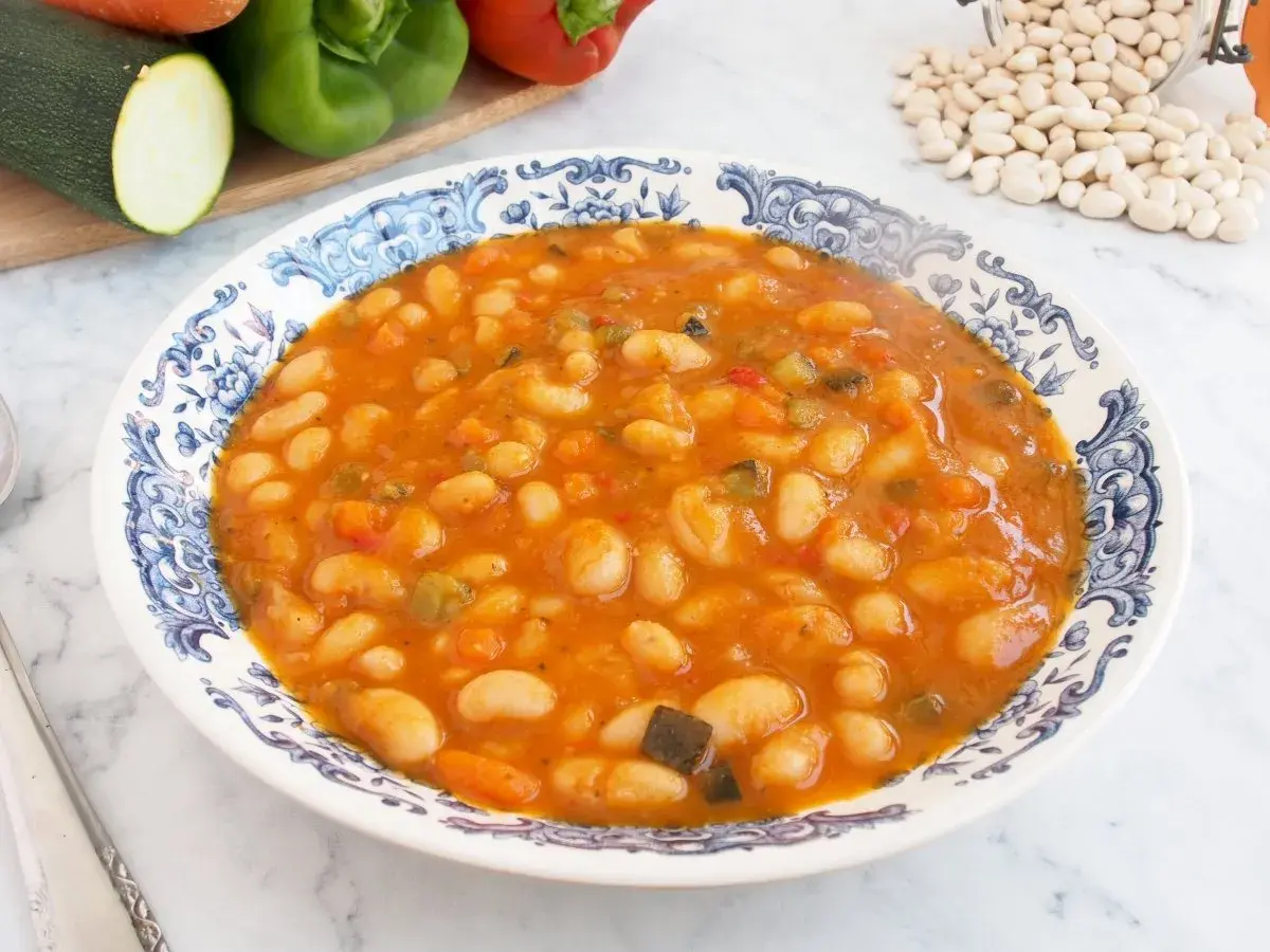 Un plato de alubias con verduras, cocinadas en una salsa rojiza y servidas en una vajilla con borde azul.