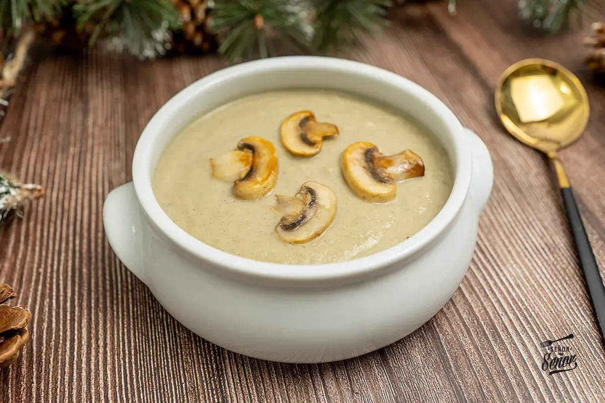 Crema de champiñones servida en un cuenco blanco, decorada con rodajas de champiñones salteados. Una cuchara dorada descansa a la derecha.