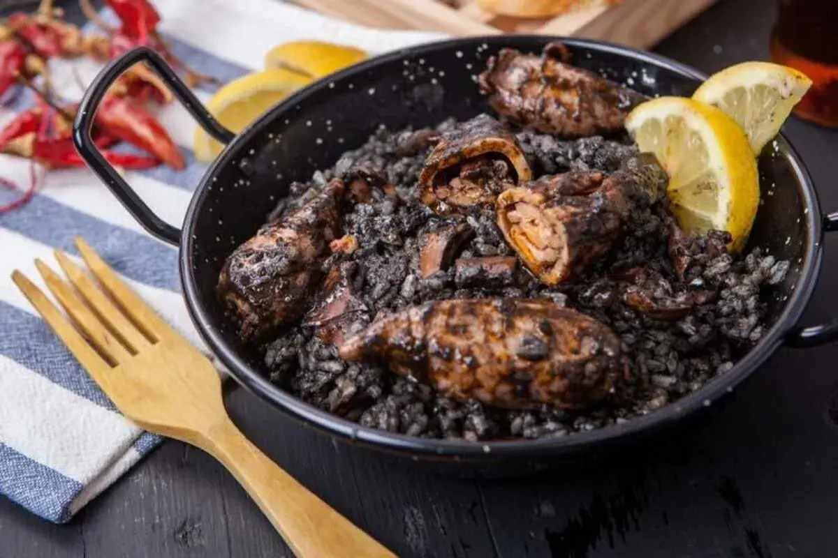 Calamares rellenos en arroz negro, servidos en una paellera con gajos de limón y una cuchara de madera.