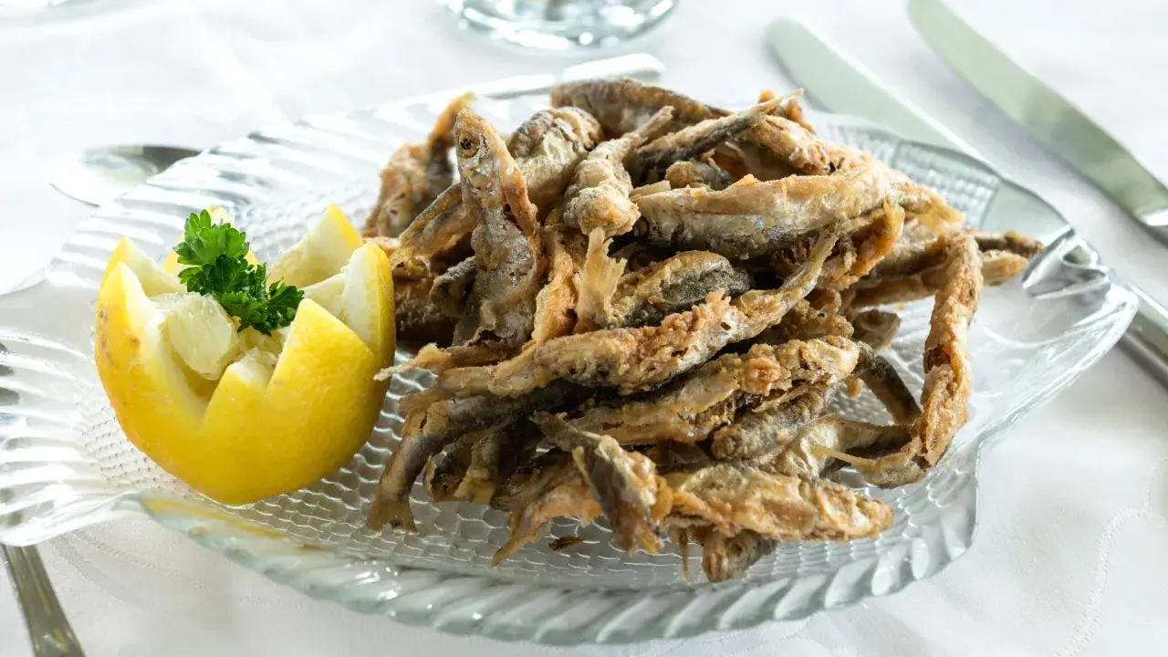 Deliciosa fritura de pescado crujiente servida en un plato de cristal con forma de pez, acompañada de un limón tallado y perejil fresco.