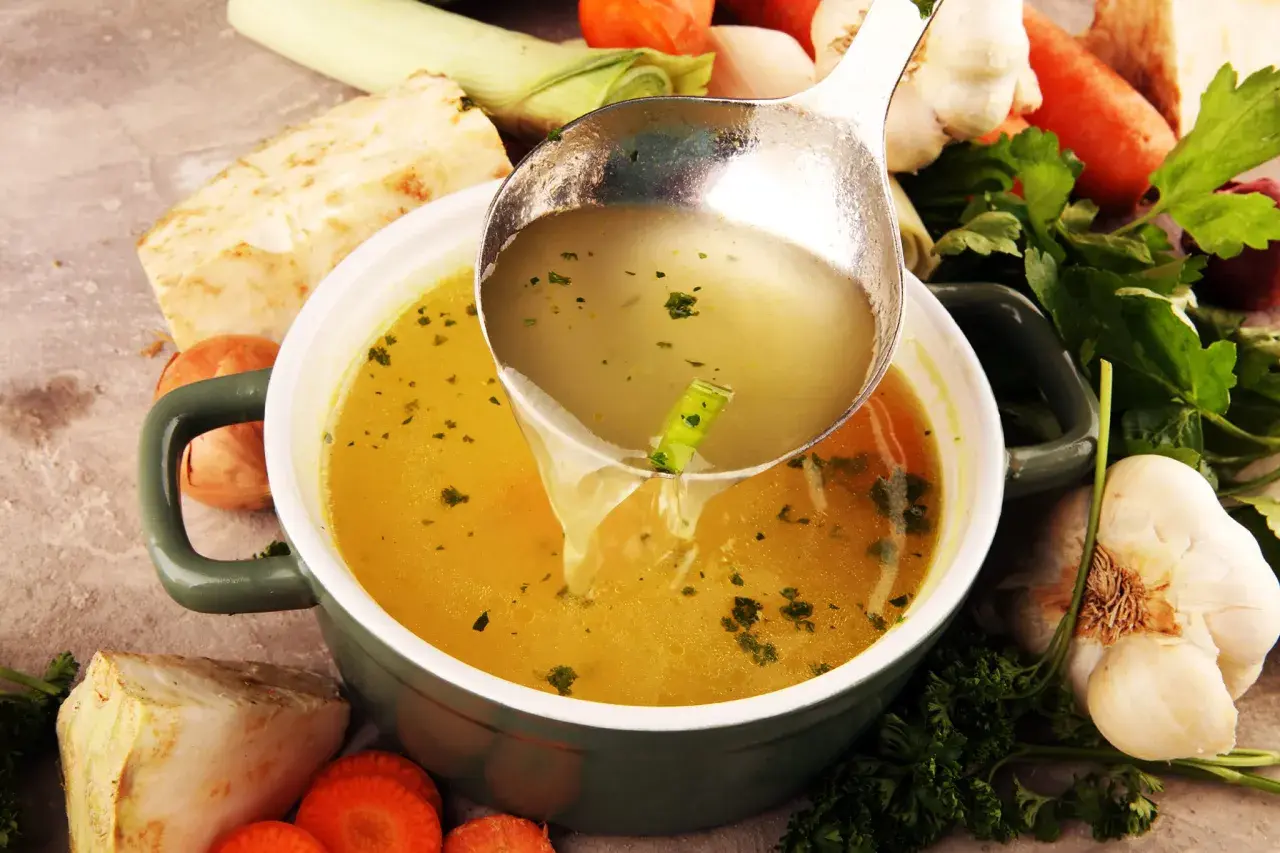 Sirviendo un reconfortante caldo de verduras recién hecho, rodeado de zanahorias, apio, puerro y ajo.