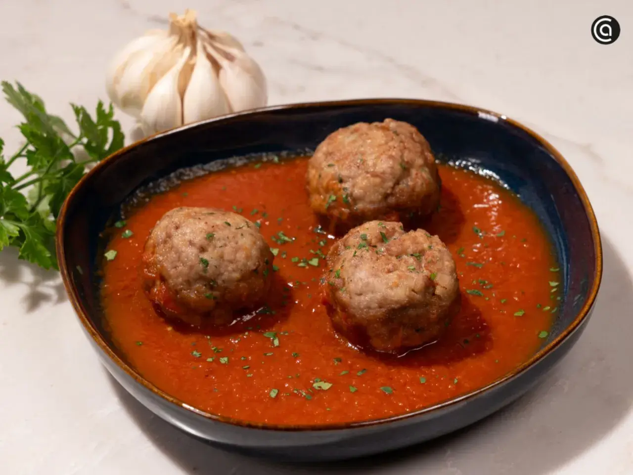 Tres albóndigas con tomate caseras, adornadas con perejil picado, junto a un ajo y perejil fresco.