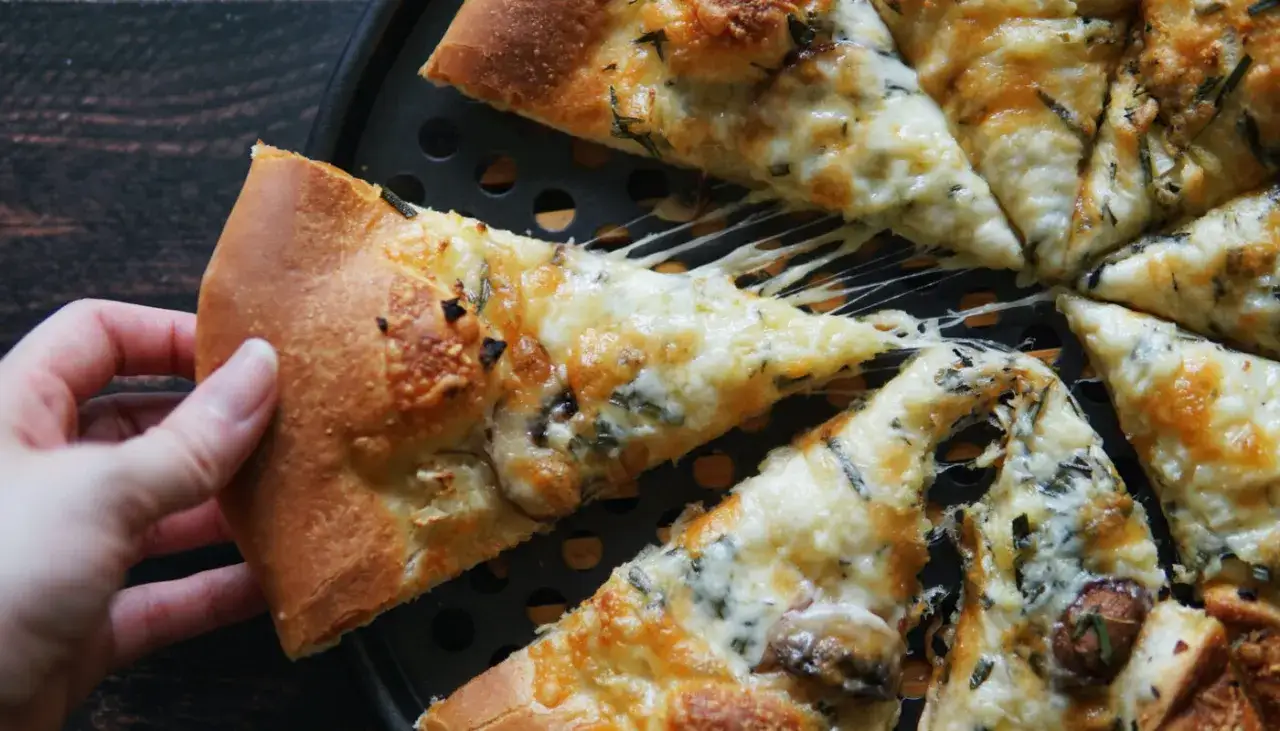 Mano toma una porción de pizza con queso derretido y hierbas. La masa pizza Thermomix luce esponjosa y dorada.