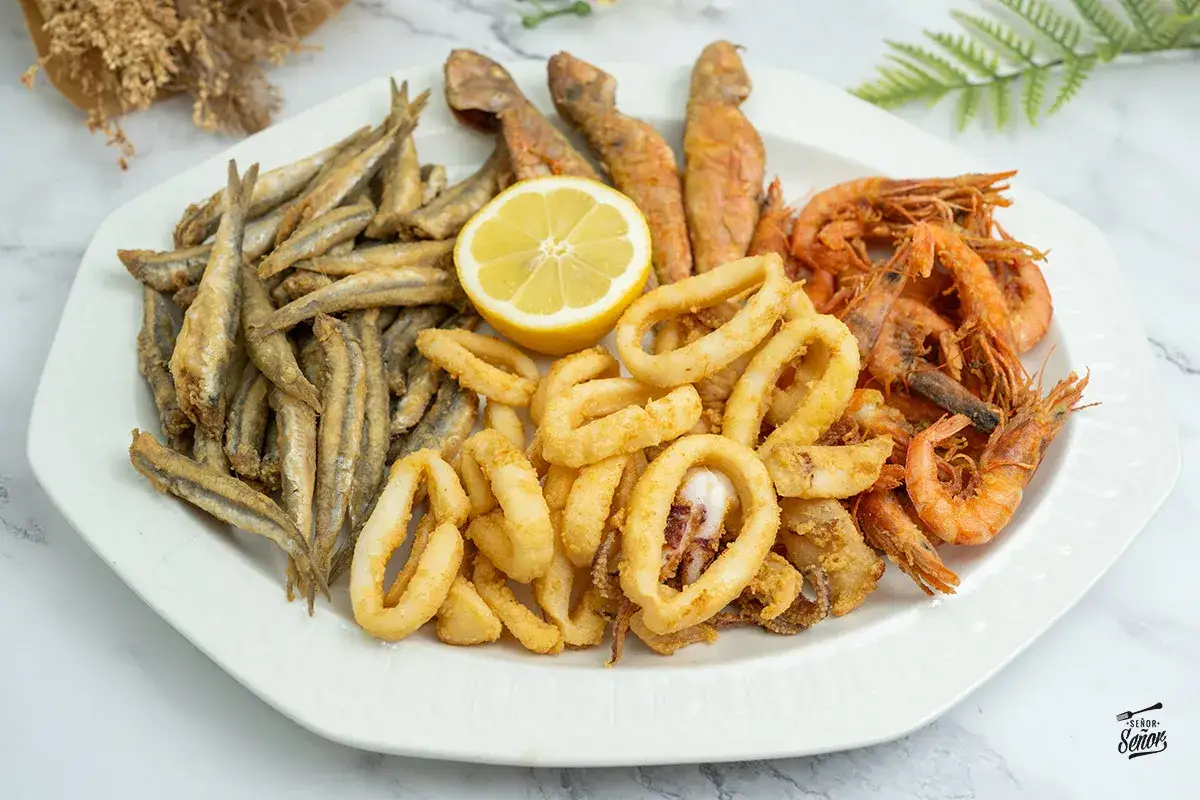 Plato de fritura de pescado con boquerones, calamares, gambas y trozos de pescado rojo, adornado con una rodaja de limón.