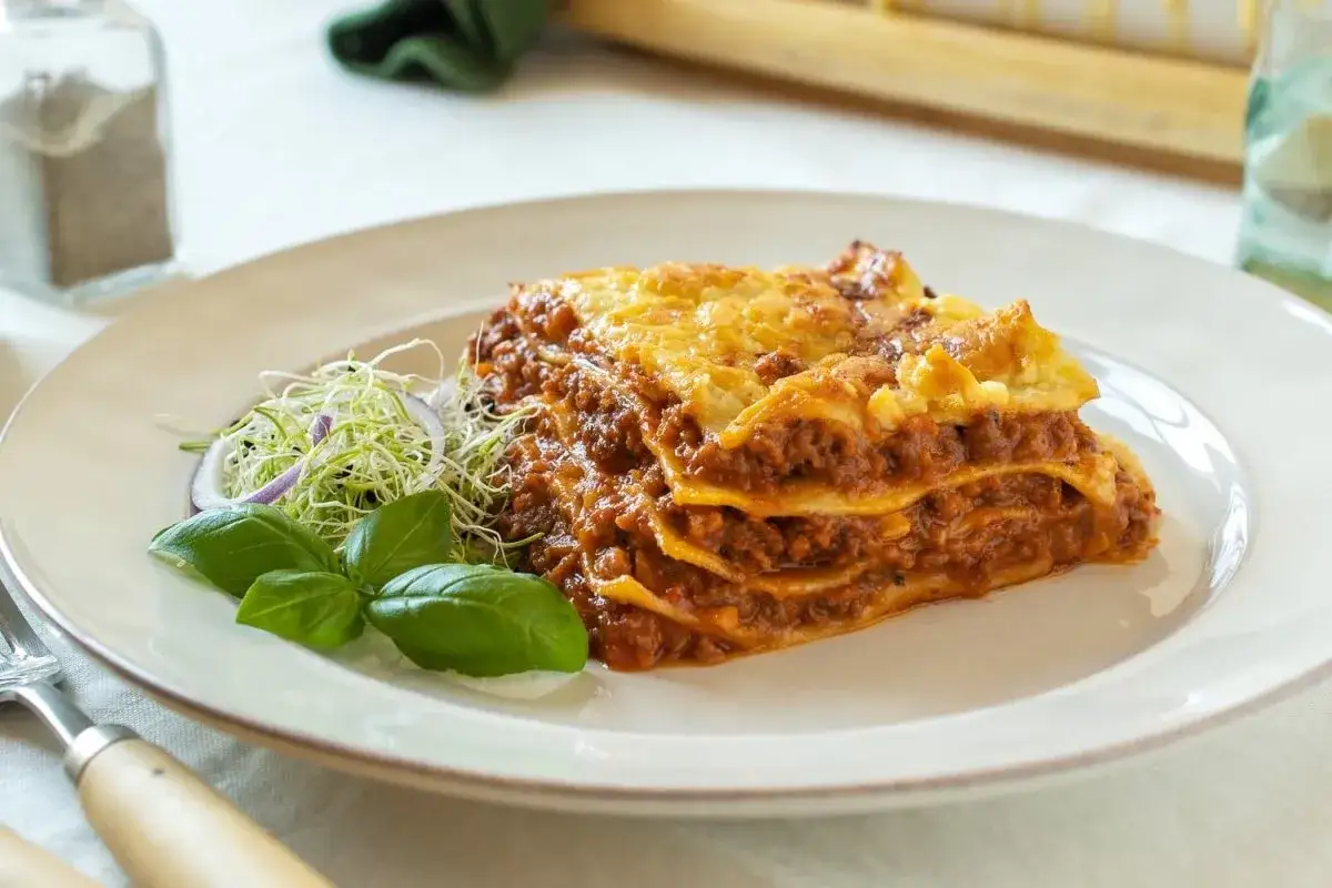 Porción de lasaña con capas de pasta, carne y queso fundido, acompañada de brotes y albahaca. Un clásico de las carnes italianas.