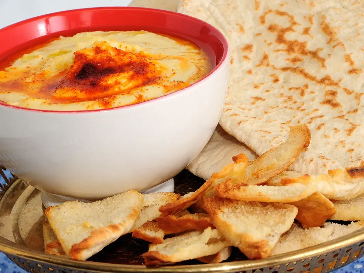 Hummus receta tradicional servido en un cuenco blanco con borde rojo, adornado con piment&oacute;n y aceite de oliva. Acompa&ntilde;ado de pan de pita y tostadas crujientes.