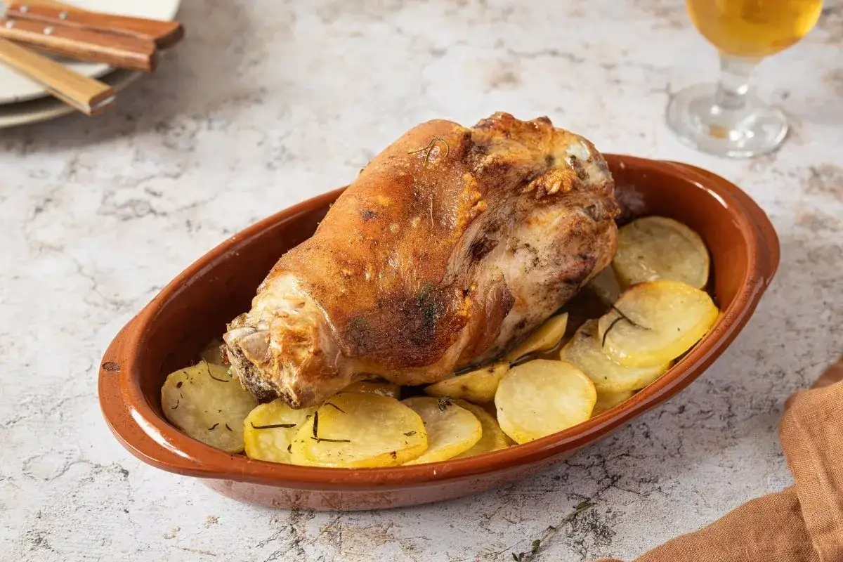 Codillo de cerdo asado, crujiente y dorado, servido con patatas en rodajas en una cazuela de barro.