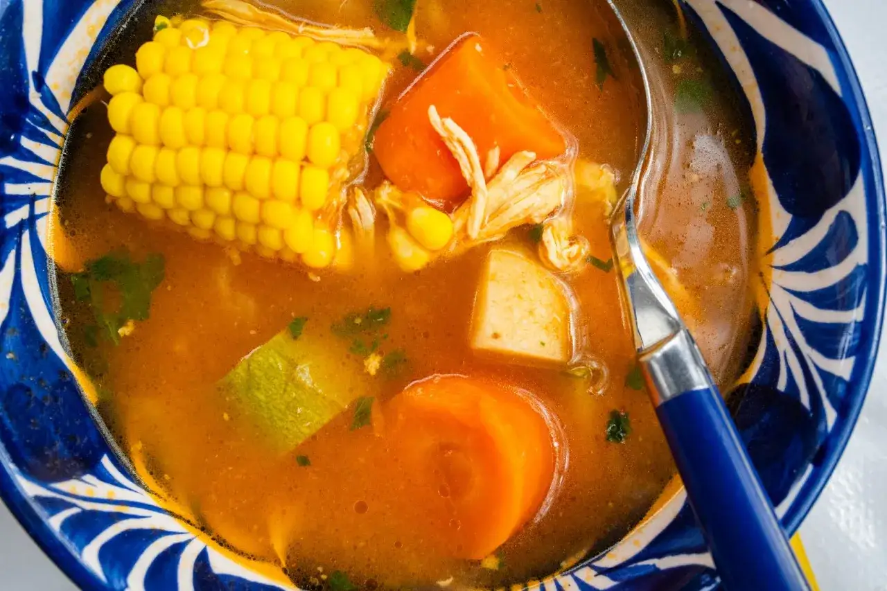 Un reconfortante caldo de pollo casero con trozos de maíz, zanahoria y patata, servido en un cuenco azul y blanco.