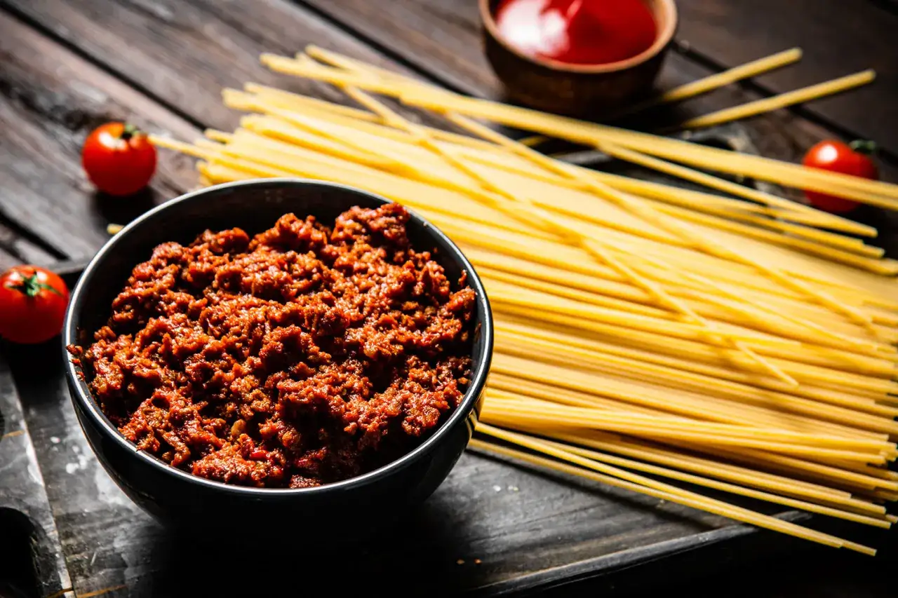 Un tazón de carne para spaghetti bolognese original, junto a tomates frescos y pasta cruda.