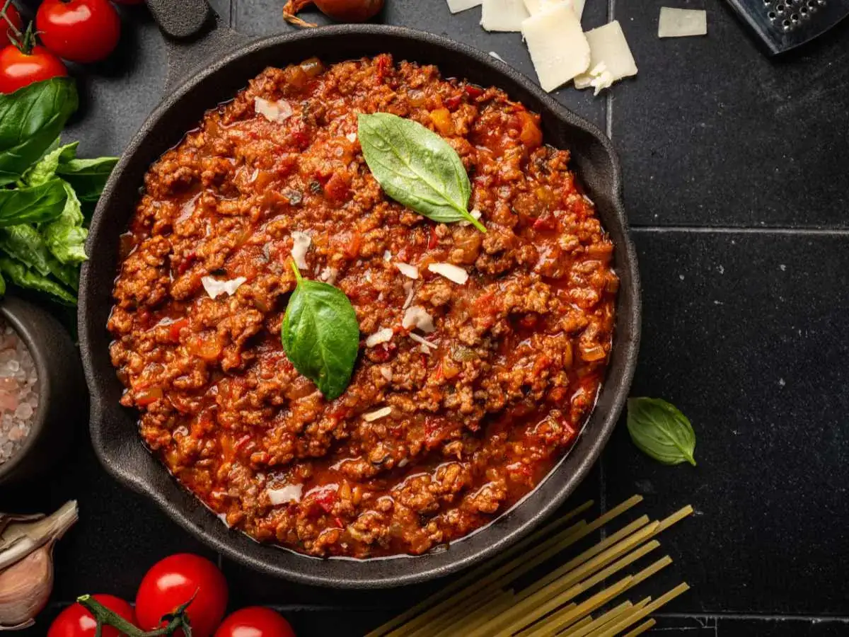 Salsa boloñesa casera en sartén de hierro fundido, con albahaca fresca, tomates y espaguetis listos para servir.
