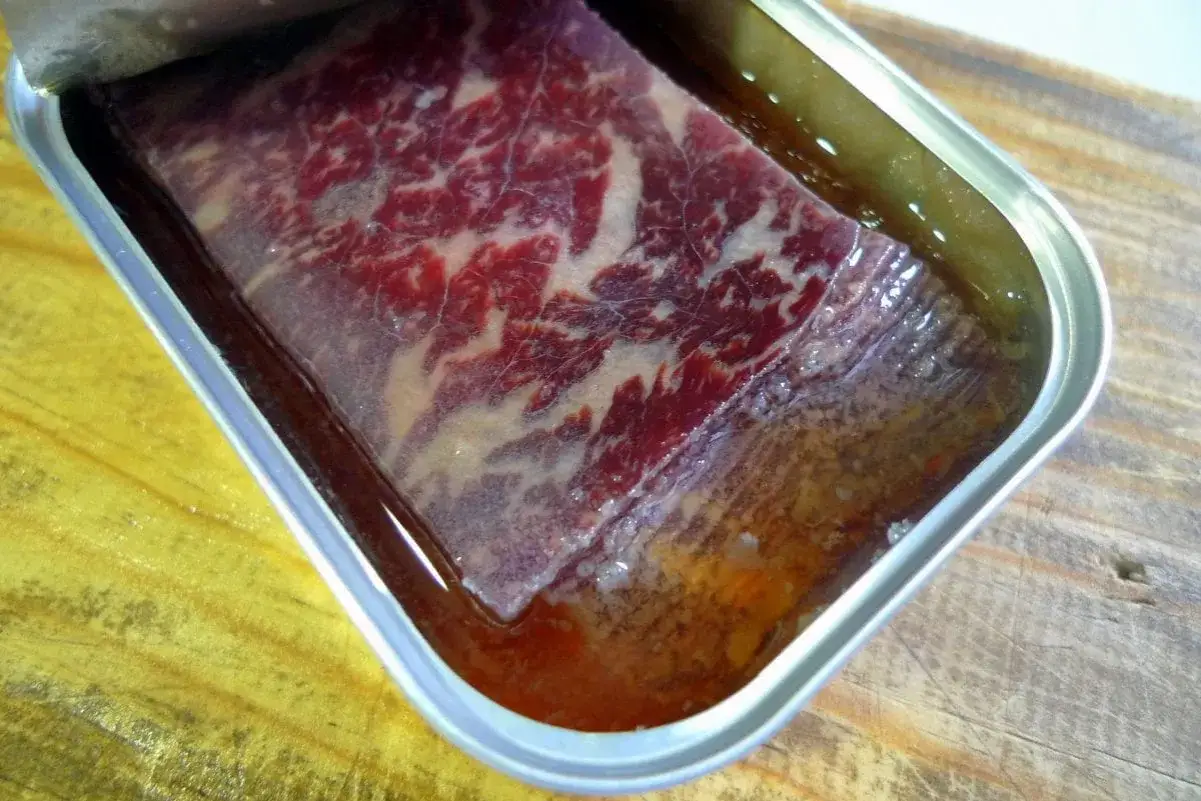 Trozo de cecina con aceite en lata, con vetas de grasa visibles, sobre una tabla de madera.